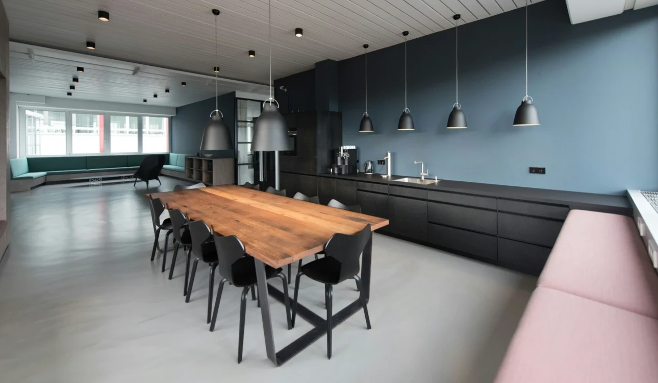 Contemporary office breakroom featuring black cabinetry, a long wooden dining table, pendant lighting, and built-in seating.
