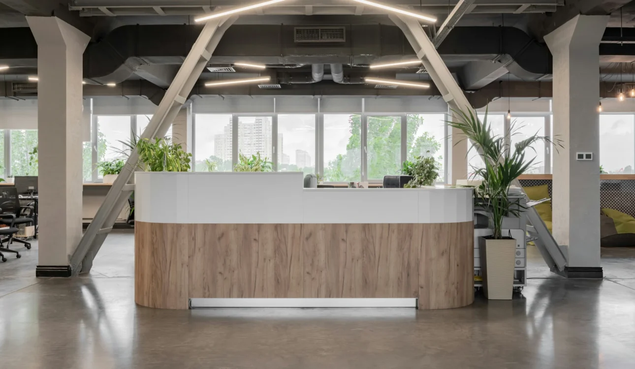 Contemporary office reception area with a wood and white desk, large windows, industrial ceiling, and green plants.