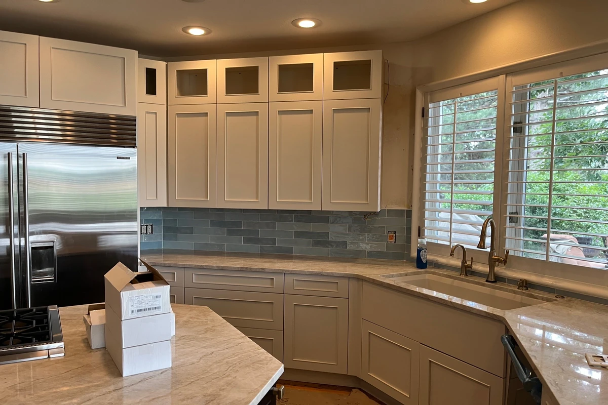 Cream kitchen cabinets with glass upper doors, blue tile backsplash, stainless refrigerator, and countertop installation in progress.