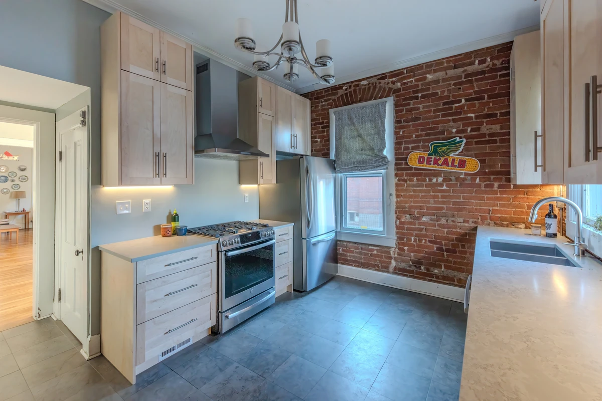 Modern kitchen with light wood cabinets, stainless steel appliances, gray tile flooring, white countertops, and an exposed brick accent wall.