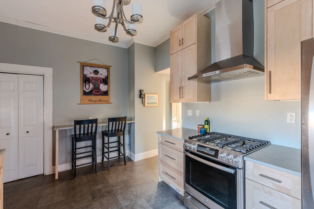 Light wood kitchen cabinets with exposed brick wall, stainless appliances, stone countertops, and modern range hood.