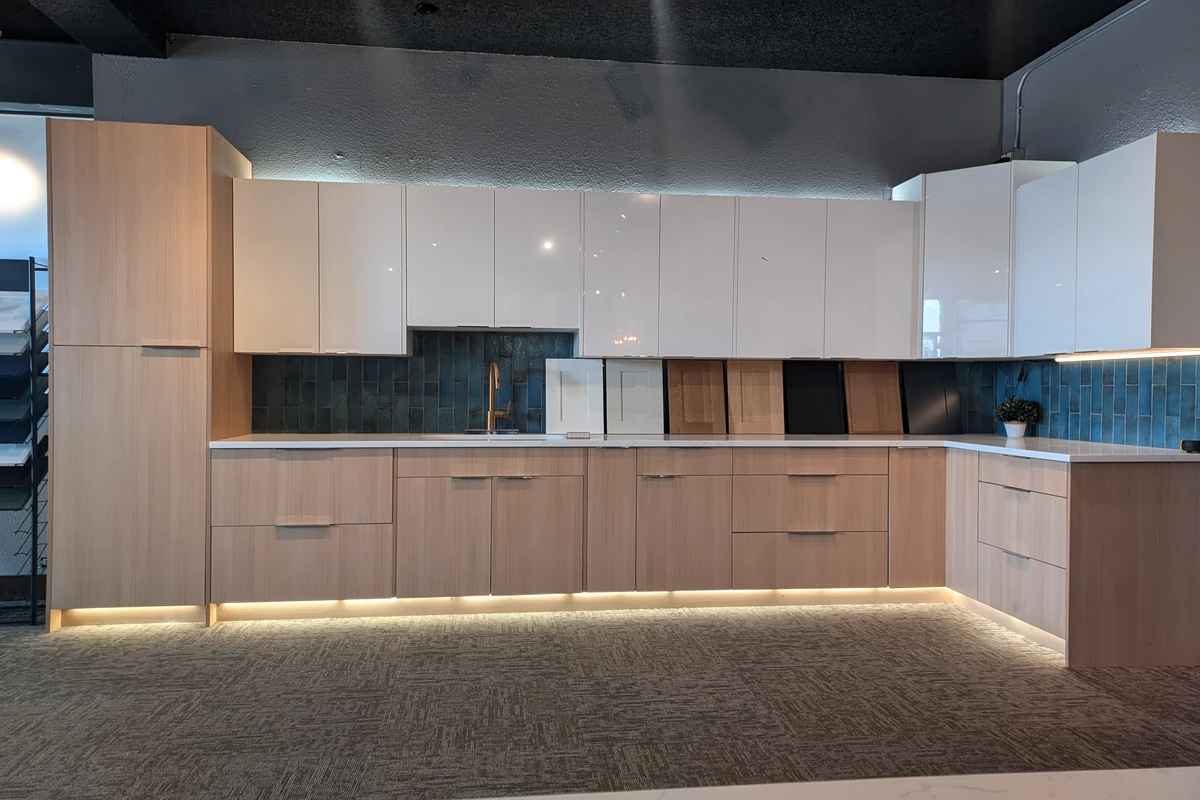 Elite Cabinetry & Granite showroom display featuring glossy white upper cabinets, light wood lower cabinets, blue tile backsplash, and integrated under-cabinet lighting.