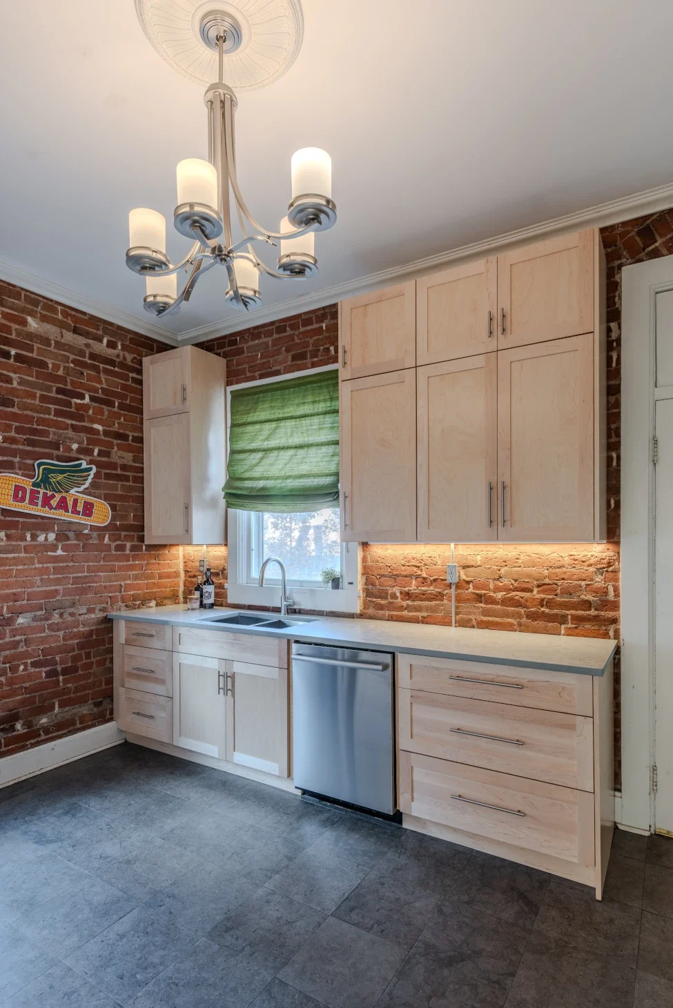 Modern kitchen with exposed brick walls, light wood cabinetry, stainless steel dishwasher, quartz countertops, and under-cabinet lighting, featured in our Elite Cabinetry company history portfolio.
